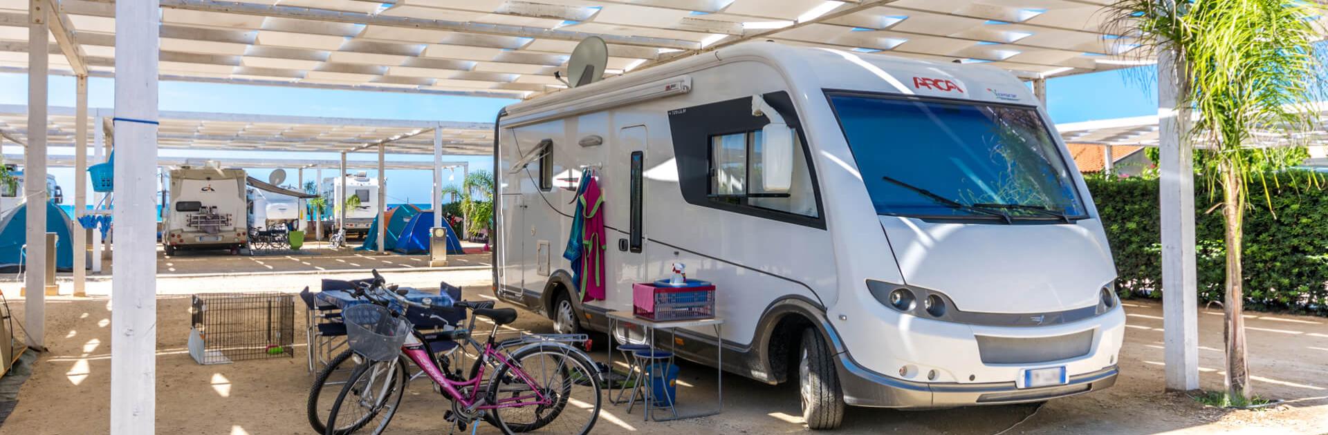 Camper parcheggiato sotto una tettoia, con biciclette e tavolo all'aperto.